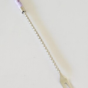 May include: A silver-toned metal cocktail fork featuring a long, twisted handle and a decorative cylindrical top section with a marbled lavender and white pattern, finished with a small, smooth silver-toned ball at the very end.
