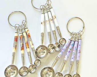 May include: Three sets of four stainless steel measuring spoons on metal rings. Each set features decorative handles with distinct patterns: one with warm earth tones, one with white and brown swirls, and one with light purple and blue floral accents.
