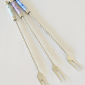 May include: Three silver-toned metal appetizer forks with long, twisted handles. Each fork features a decorative cylindrical handle end with a marbled pattern in light blue, lavender, and dark blue, topped with a small polished metal sphere.