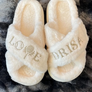 May include: A pair of cream-colored, fluffy slippers with a criss-cross design. The left slipper has the word "LOVE" and a tennis racket design, while the right slipper says "DRISA" in gold lettering. The slippers are on a gray faux fur surface.