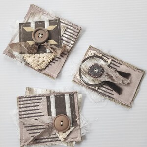 May include: Three handmade fabric embellishments with brown and white patterns and brown buttons. The embellishments are made with different textures and fabrics, including lace and burlap.