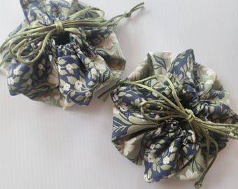 May include: Two round fabric pouches featuring a floral pattern in shades of sage green, navy blue, and white. Each pouch has a gathered top secured with a thin olive green drawstring cord tied in a bow, creating a ruffled edge around the opening.