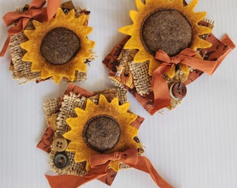 May include: Three handmade sunflower decorations. Each has a yellow felt flower with a brown center, set on a square of burlap and patterned fabric. They are adorned with orange ribbons and buttons, creating a rustic, autumnal theme.