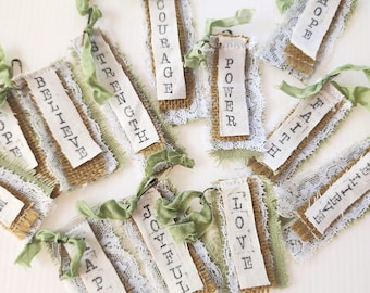 May include: A collection of rectangular fabric tags featuring frayed burlap, white lace trim, and sage green ribbon ties. Each tag displays a single word stamped in black ink, including Believe, Strength, Courage, Power, Hope, Faith, Happy, Joyful, and Love.