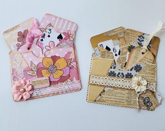 May include: Two decorative paper pockets containing playing cards and tags. The left pocket is pink with floral patterns, lace, and a fabric flower. The right pocket features vintage dictionary page text, black floral patterns, lace, and a fabric flower.