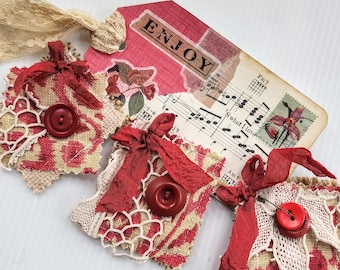 May include: Three red and white fabric ornaments with red bows and buttons. The ornaments are decorated with lace and have a rustic, vintage look. The ornaments are arranged on a white surface with a sheet of music in the background. The sheet music has the word "Enjoy" written on it.