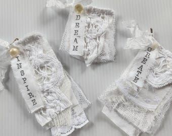May include: Three white fabric tags featuring layered lace and textile scraps. Each tag is topped with a small cream-colored bead and a tied ribbon. The tags are printed with the words INSPIRE, DREAM, and CREATE in a black typewriter-style font.