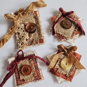 May include: Four handmade fabric embellishments. Each square features a button, burlap, and patterned fabric in shades of brown, beige, and burgundy. They are adorned with ribbons and decorative stitching, perfect for scrapbooking or crafting projects.