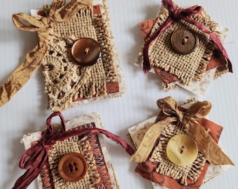May include: Four handmade fabric embellishments. Each square features a button, burlap, and patterned fabric in shades of brown, beige, and burgundy. They are adorned with ribbons and decorative stitching, perfect for scrapbooking or crafting projects.