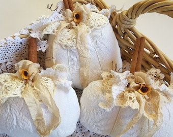 May include: Three white fabric pumpkins with cinnamon stick stems, lace accents, and small yellow flower embellishments. The pumpkins are arranged in a woven basket, creating a rustic, fall-themed display.