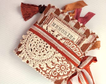 May include: A square fabric-covered book with a brown and white checkered pattern, featuring a cream-colored crochet doily, a white lace trim, and a fabric label with the word MEMORIES. The book is accented with a brown tassel and ribbons.