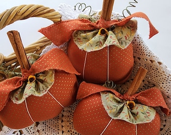 May include: Three handmade fabric pumpkins with cinnamon stick stems, orange polka dot bodies, and decorative bows. The bows are made of orange and green patterned fabric. The pumpkins are arranged in a wicker basket, suggesting a fall or harvest theme.
