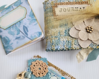 May include: A small square notebook with a blue floral cover and a blank label, a rectangular fabric-covered journal with the word JOURNAL printed on a burlap strip, and a small rectangular fabric tag featuring lace and a crocheted flower.