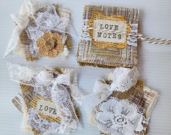 May include: Four square burlap and lace embellishments featuring frayed edges, white lace bows, and fabric flowers. One piece displays the text LOVE NOTES on a dictionary page cutout, another shows the word LOVE, and all include twine accents.