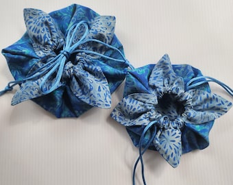 May include: Two round fabric drawstring pouches in varying sizes, featuring a blue and teal batik-style pattern with light blue teardrop and leaf motifs. Each pouch has a gathered top secured by matching blue cord drawstrings.