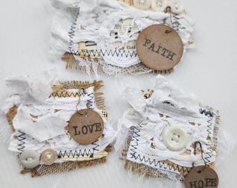 May include: Three rectangular fabric embellishments featuring layered white lace, frayed burlap, and paper scraps with musical notes. Each piece includes white buttons and a round brown tag attached with a safety pin, displaying the words FAITH, LOVE, or HOPE.
