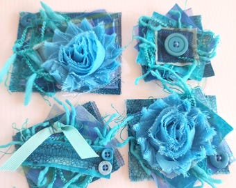 May include: Four handmade textile art pieces in shades of blue and turquoise. Each square features layered fabric, decorative stitching, and embellishments like buttons, ribbons, and fabric flowers. The pieces have a textured, artistic appearance.