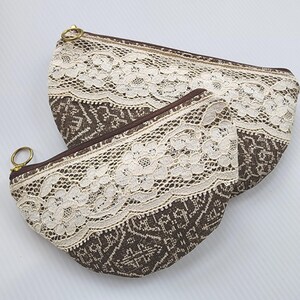 May include: Two brown fabric pouches with white lace trim. The pouches are lined with a brown and white geometric pattern. The pouches are closed with a zipper.