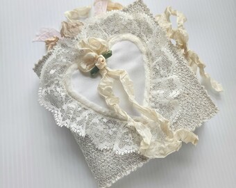 May include: A handmade, heart-shaped decorative item. The item features a white heart shape, trimmed with delicate lace and adorned with a cream-colored flower and ribbons. The base is a textured gray fabric, with additional cream and pink ribbons.