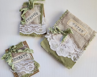 May include: Three rectangular fabric-covered items featuring sage green and tan burlap, white floral lace trim, and sage green ribbon bows. Each item has a white fabric label with black text reading INSPIRE, CREATE, or NOTES respectively.