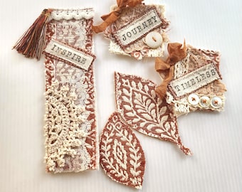 May include: A collection of fabric items including a rectangular bookmark with a tassel and the word INSPIRE, two square patches with the words JOURNEY and TIMELESS, and two leaf-shaped fabric pieces, all featuring brown and cream lace patterns.
