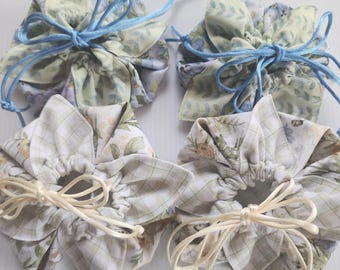 May include: Four floral fabric drawstring pouches. Two pouches have blue drawstrings, and two have cream-colored drawstrings. The fabric features a pattern of flowers and leaves in shades of green, yellow, and white. The pouches are gathered at the top.