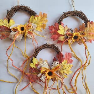 May include: Three autumn-themed wreaths made of woven twigs, adorned with colorful faux leaves in shades of red, yellow, and orange. Each wreath features a sunflower, burlap and lace bow, and long, flowing ribbons.