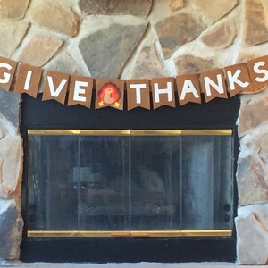 May include: A brown paper banner with white letters that reads "GIVE THANKS" with a small turkey illustration in the middle. The banner is hanging above a fireplace.