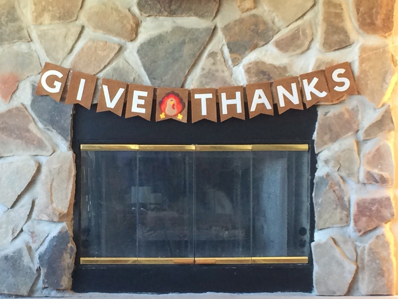 May include: A brown paper banner with white letters that reads "GIVE THANKS" with a small turkey illustration in the middle. The banner is hanging above a fireplace.
