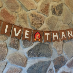 May include: A brown felt banner with white letters spelling "GIVE THANKS" and a small felt turkey in the middle. The banner is hanging on a stone wall.
