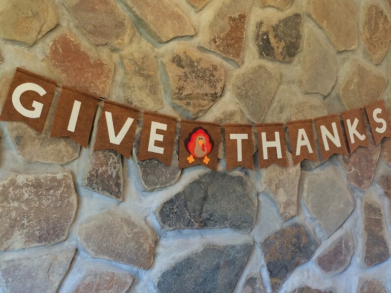 May include: A brown felt banner with white letters spelling "GIVE THANKS" and a small felt turkey in the middle. The banner is hanging on a stone wall.