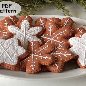 Puede incluir: Un plato de adornos de copo de nieve de jengibre de fieltro. Los copos de nieve son marrones con bordados blancos y algunos son blancos con bordados marrones.