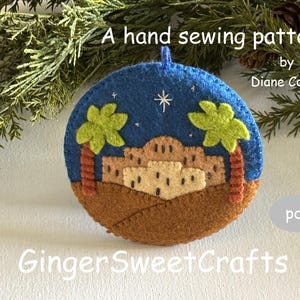 May include: A hand-sewn felt ornament with a nativity scene design. The ornament features a blue sky with a star, palm trees, and a tan-colored town. The text "A hand sewing pattern" and "GingerSweetCrafts" are visible.