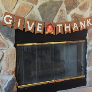 May include: A brown felt banner with white letters that reads "GIVE THANKS" with a small felt turkey in the middle. The banner is hanging above a black fireplace with a glass door.