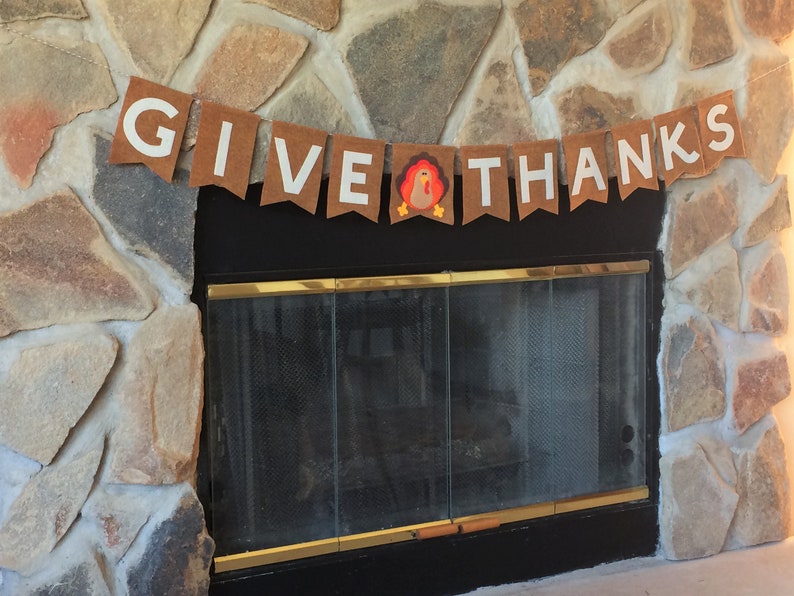 May include: A brown felt banner with white letters that reads "GIVE THANKS" with a small felt turkey in the middle. The banner is hanging above a black fireplace with a glass door.