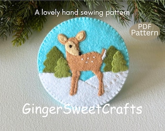 May include: A circular felt craft project featuring a brown fawn with white spots standing in a snowy landscape with green pine trees against a bright blue sky. The text A lovely hand sewing pattern, PDF Pattern, and GingerSweetCrafts are visible.
