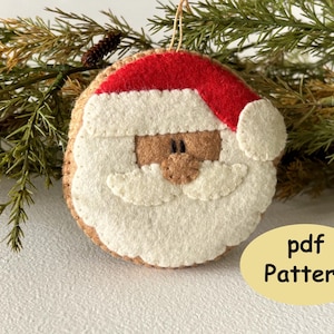 May include: Felt ornament of Santa Claus with a white beard and red hat. The ornament has a brown face and black eyes. The text "pdf Pattern" is visible in the image.