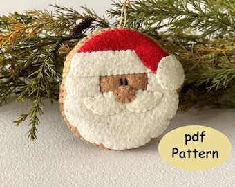 May include: A round felt Santa Claus ornament featuring a red hat with a white brim, a tan face, and a white beard. The ornament has visible stitching along the edges and includes a hanging loop. A yellow oval graphic displays the text pdf Pattern.
