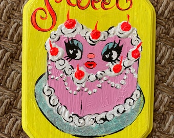 Op de afbeelding: Handgeschilderd, achthoekig houten bord met een gele achtergrond. Het bord toont een roze cake met een gezicht, gegarneerd met kersen en witte glazuur. Het woord "Sweet" staat in rode letters boven de cake.