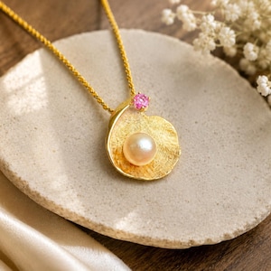 May include: A gold necklace featuring a textured, shell-shaped pendant with a central cream-colored pearl and a small, round, bright pink gemstone accent at the top. The pendant hangs from a delicate gold chain, showcasing a polished metal finish.