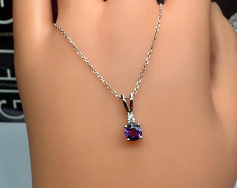 Può includere: Una collana d'argento con un piccolo ciondolo con una gemma rotonda, viola scuro e una pietra più piccola e trasparente. Il ciondolo pende da una delicata catena. La collana è esposta su una mano.