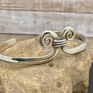 Op de afbeelding: Een zilveren armband met een uniek, gedraaid ontwerp. De armband is gemaakt van dik, glad metaal en heeft een eenvoudige, elegante uitstraling.
