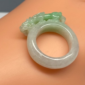 Puede incluir: Un anillo de jade con un diseño de dragón tallado. El anillo es de color verde pálido con un centro blanco.