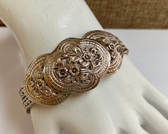 May include: Silver-toned bracelet featuring three large, ornate oval links with floral and leaf designs. The bracelet has a textured, woven band and is displayed on a white surface.