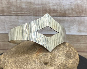 May include: A silver bracelet with a textured, hammered finish and a diamond-shaped cutout. The bracelet is curved and has a modern, minimalist design.