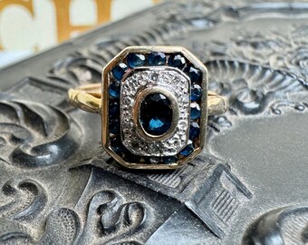 Puede incluir: Un anillo de oro con una cara rectangular. La cara presenta una piedra central ovalada de color azul oscuro, rodeada de pequeñas piedras blancas y una fila exterior de piedras de color azul oscuro. El anillo está sobre un fondo texturizado.
