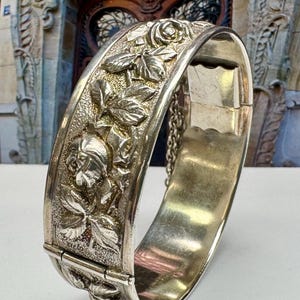 May include: A silver hinged bangle bracelet with a detailed floral design featuring roses and leaves.