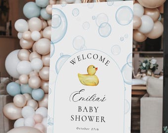 May include: A baby shower welcome sign on a wooden easel. The sign is white with blue bubble graphics and the words "Welcome" and "Emilia's Baby Shower" with a yellow rubber duck illustration. Balloons in shades of white, beige, and blue are in the background.