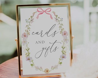 May include: A gold-framed sign with a white background and a floral wreath design. The sign says "cards and gifts" in a cursive font with a pink ribbon bow at the top.