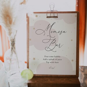 May include: A sign with the text "Mimosa Bar" in cursive script, with a pink and white background and gold stars. The sign also includes instructions for making a mimosa: "Pour some bubbly, add a splash of juice, top with fruit, cheers!"